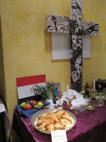 Weltgebetstag Auf einem Tisch ist ein Buffet mit landesspezifischen Köstlichkeiten aufgebaut, dahinter ein Kreuz und eine Landesflagge
