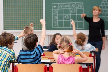 Schulen und Hochschulen In einem Klassenzimmer sitzen Kinder, melden sich und warten, dass die Lehrerin, die an der Tafel steht, sie aufruft